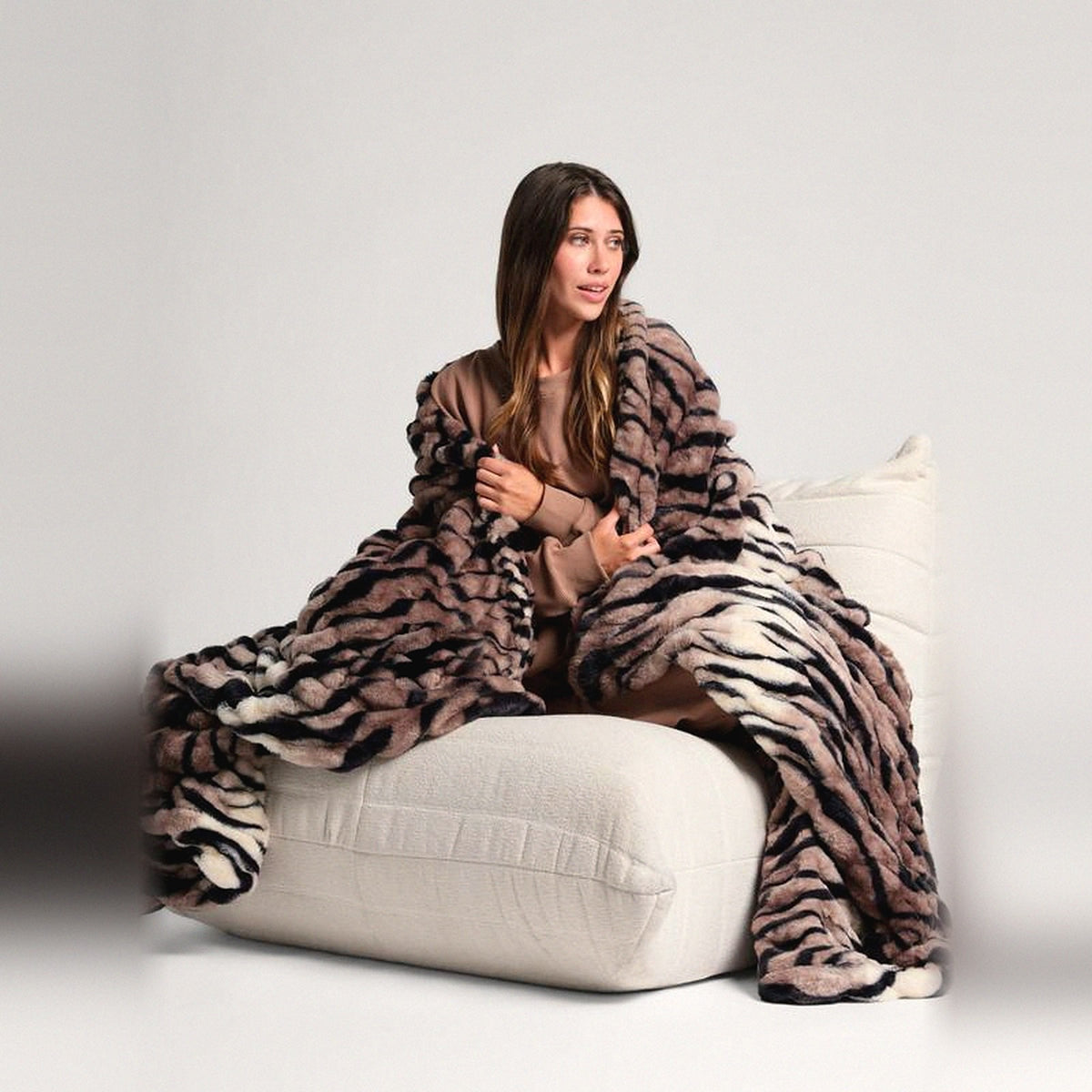 Mochi Mellow Cloud Throw in Brown Tiger — Brown tiger-print ruched faux-fur throw wrapped around a woman on a cream lounger