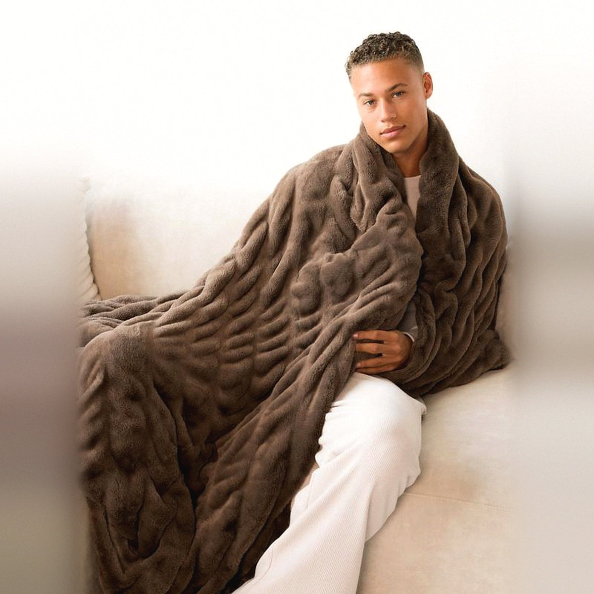 Mochi Mellow Cloud Throw in Mocha — Mocha brown ruched faux-fur throw wrapped around a man sitting on a cream sofa