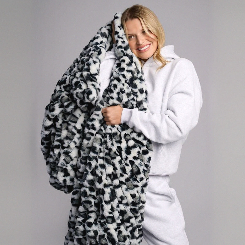 Mochi Mellow Cloud Throw in Snow Leopard — Snow leopard-print ruched faux-fur throw held up around a woman's shoulders