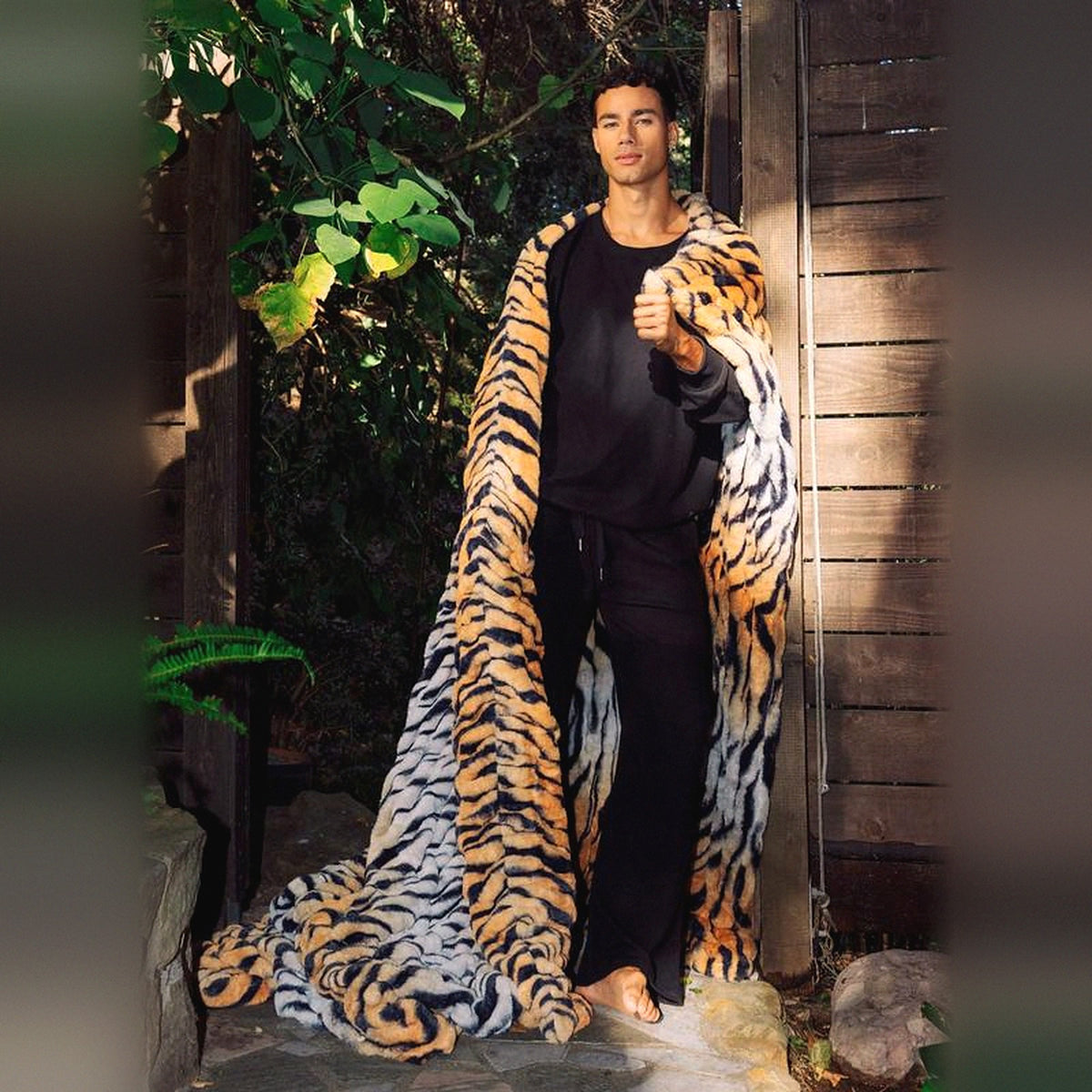 Mochi Mellow Cloud Throw in Tiger — Tiger-print ruched faux-fur throw draped over a man's shoulders in a garden